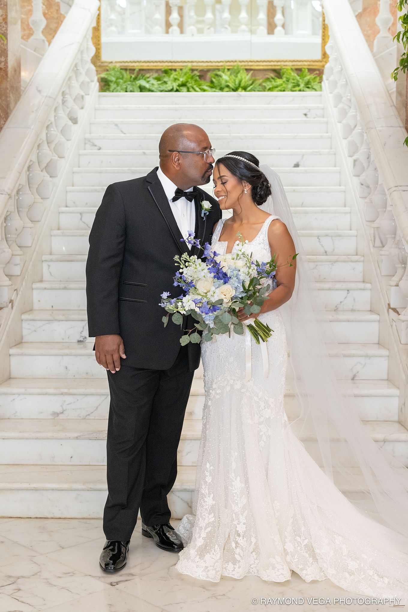 Bride and groom share a romantic moment on marble staircase, bride holding blue and white bouquet in elegant lace gown.