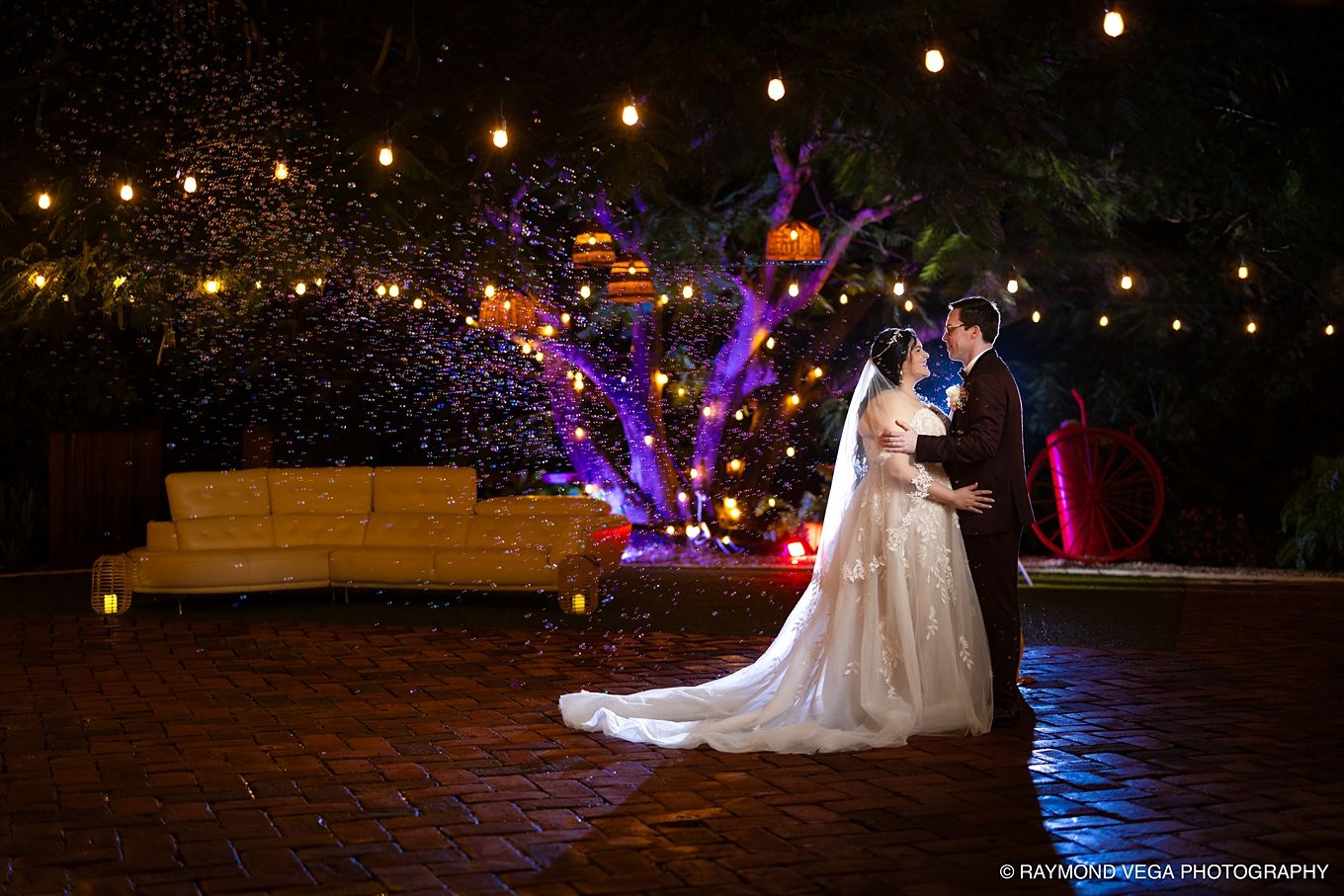 Bride and groom share romantic moment outdoors at night, surrounded by glowing lights and colorful tree illuminations.
