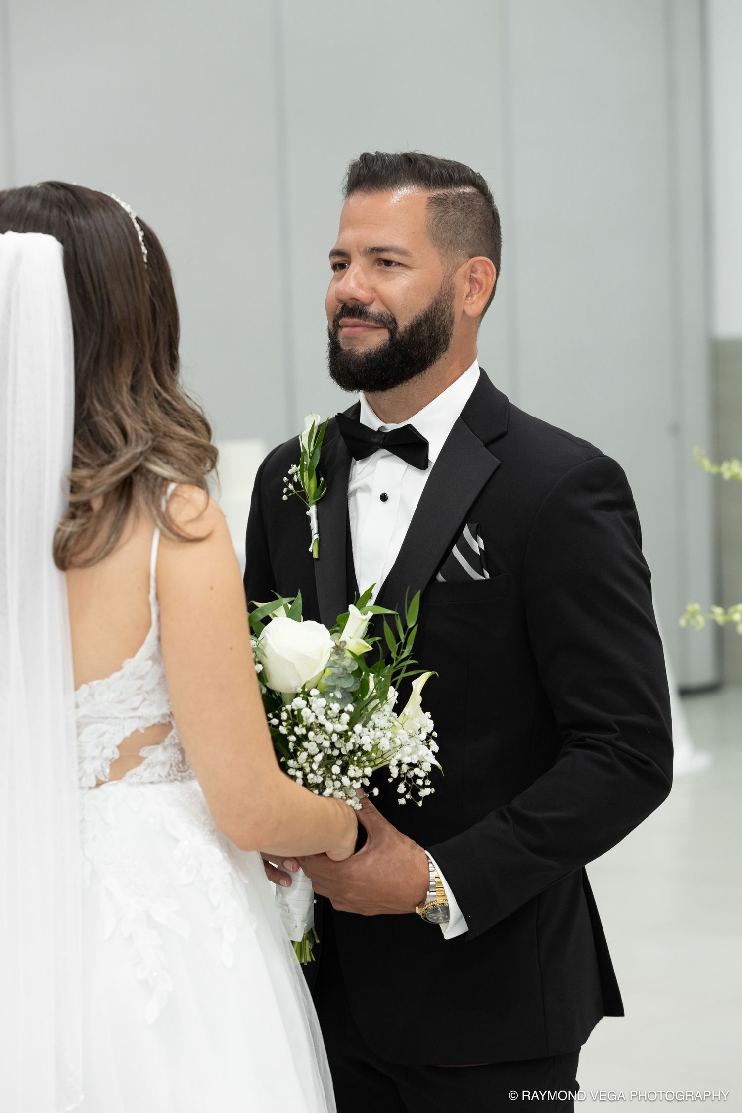 Groom in black tuxedo smiling at bride during wedding ceremony while holding white floral bouquet.