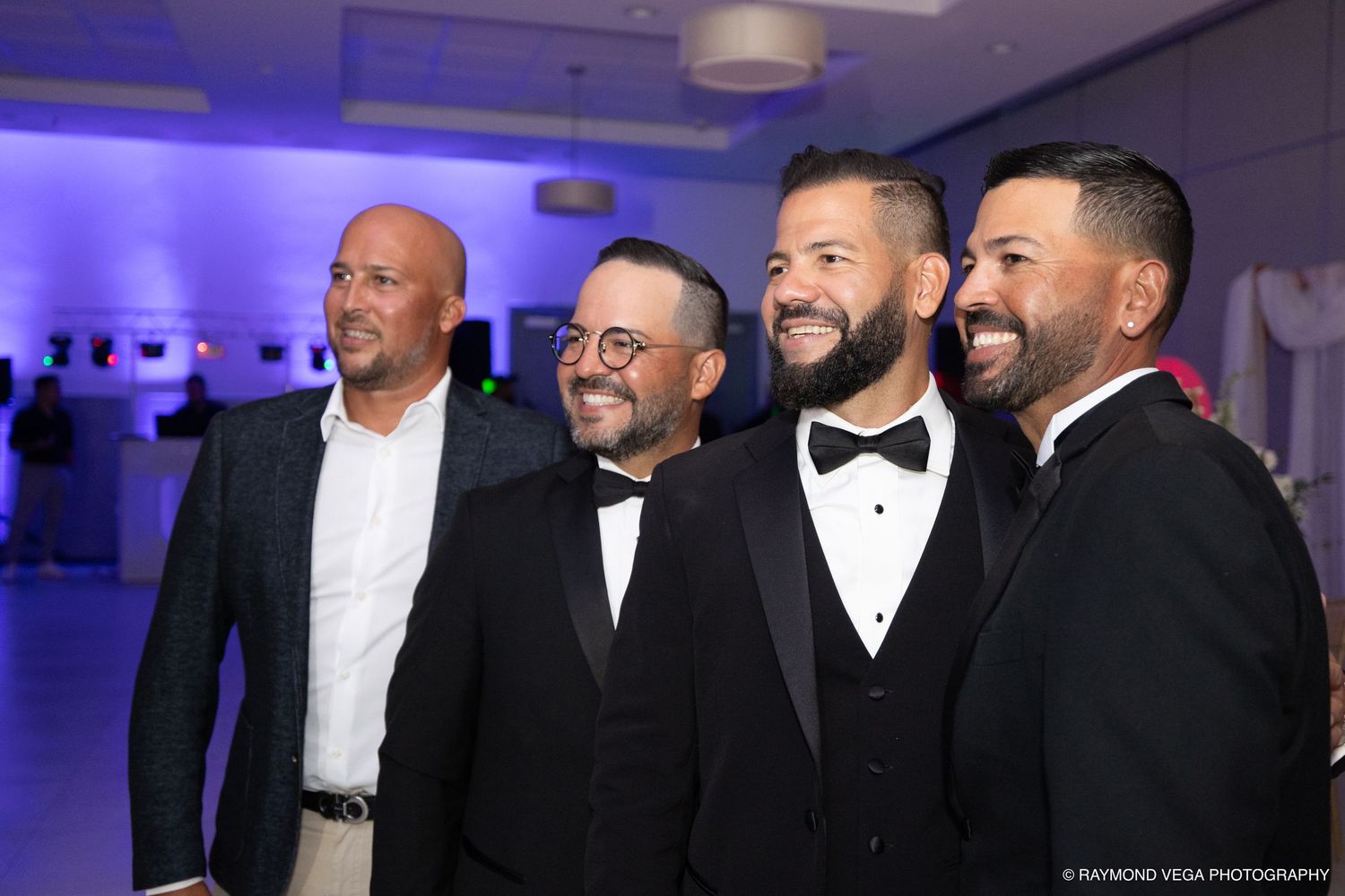 Four men in formal attire smile together at an elegant event with blue ambient lighting.