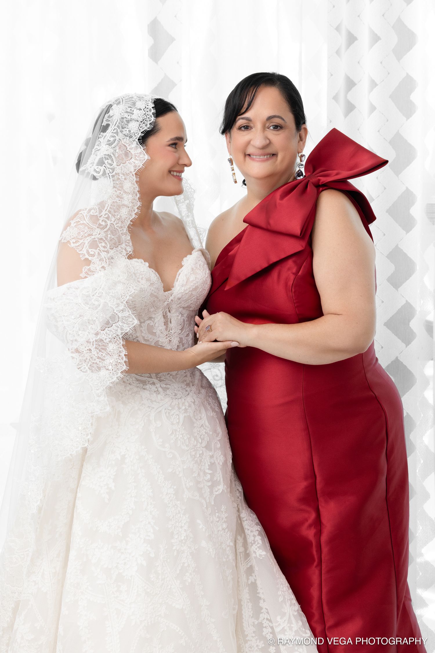 Bride in white lace gown and veil poses with woman in red satin bow dress at wedding celebration.