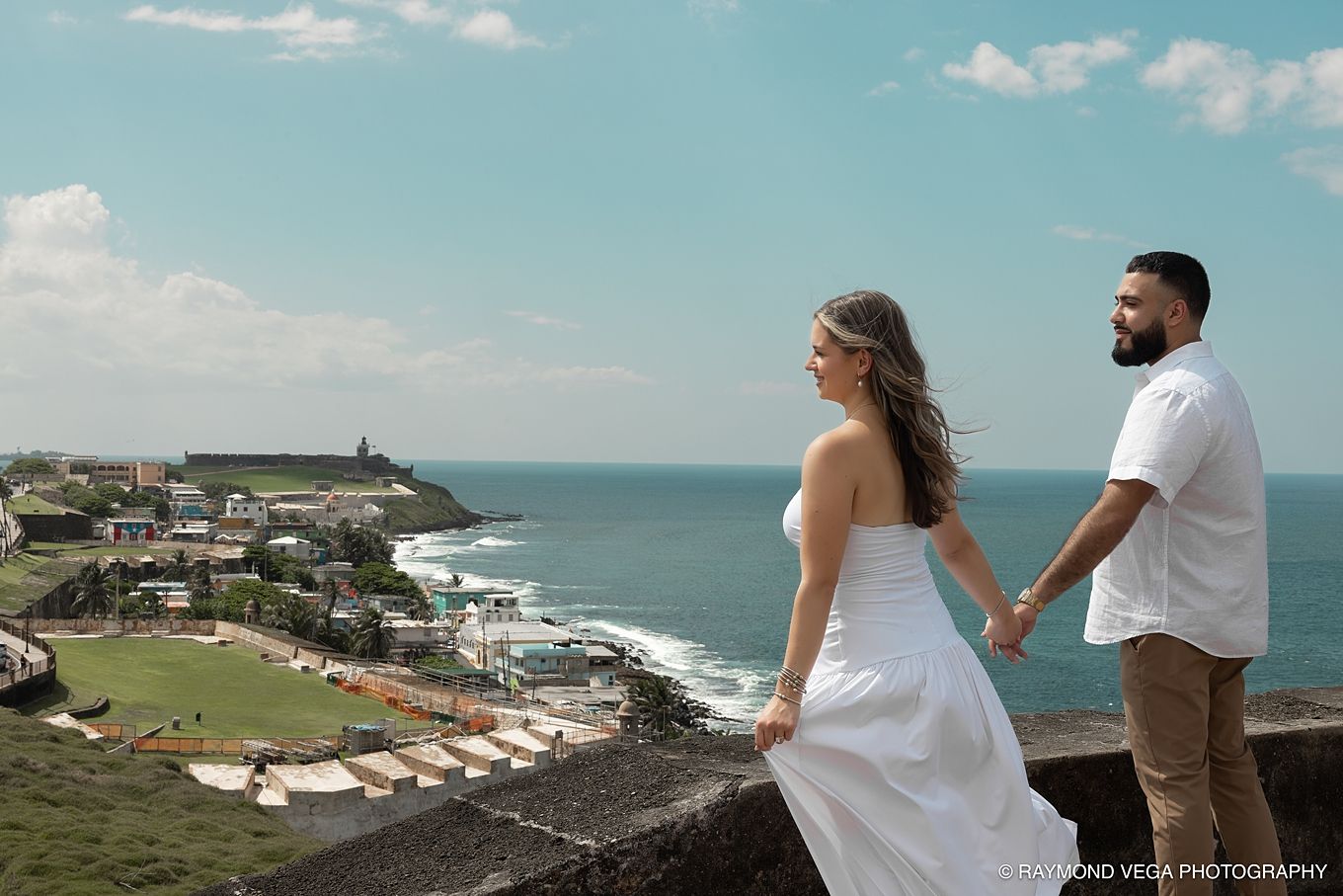 Couple in wedding attire holding hands overlooking Old San Juan coastline and El Morro fortress on a sunny day.