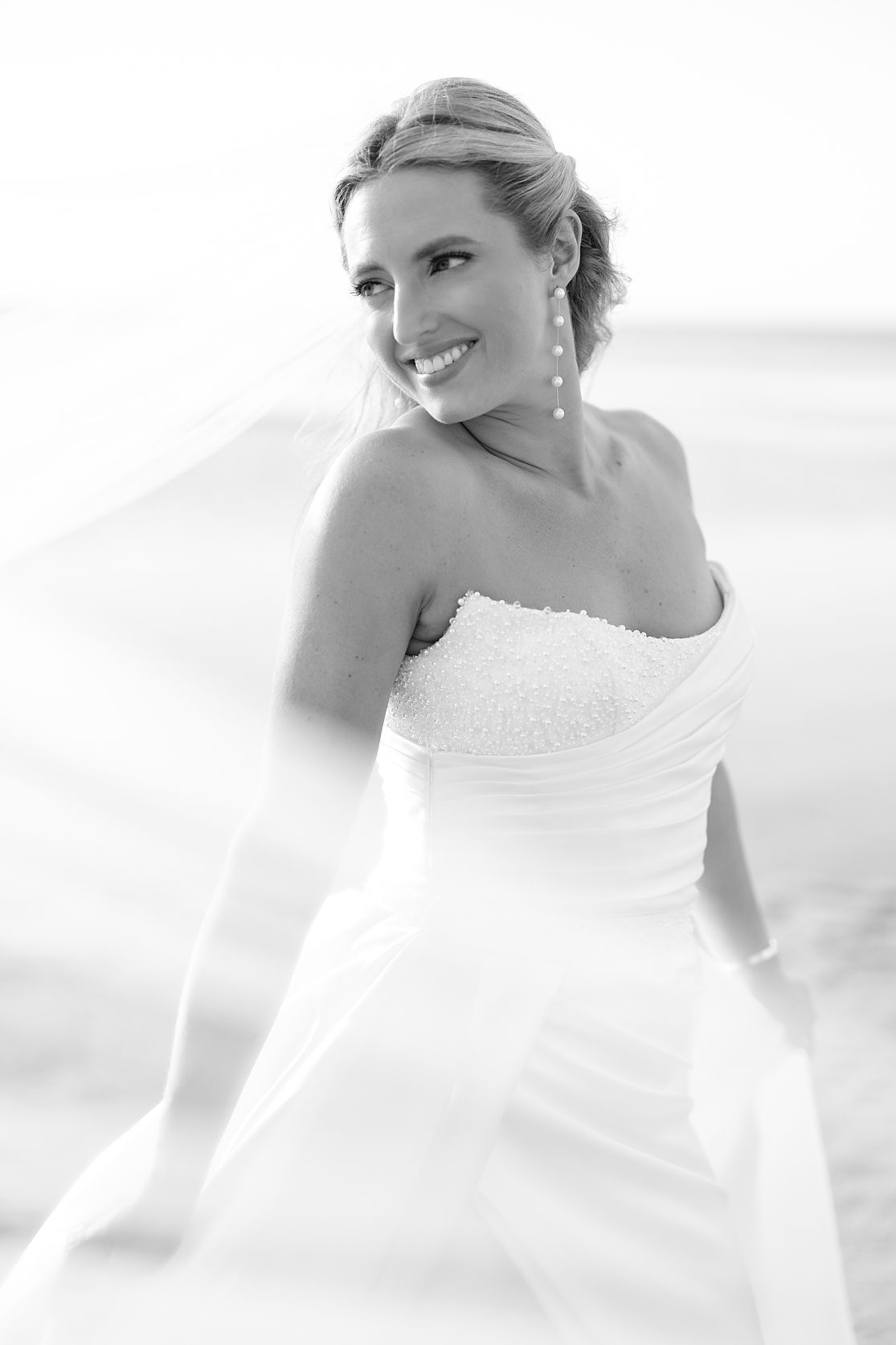 Smiling bride in strapless white gown with veil flowing in breeze, black and white outdoor wedding portrait. Raymond Vega Photography