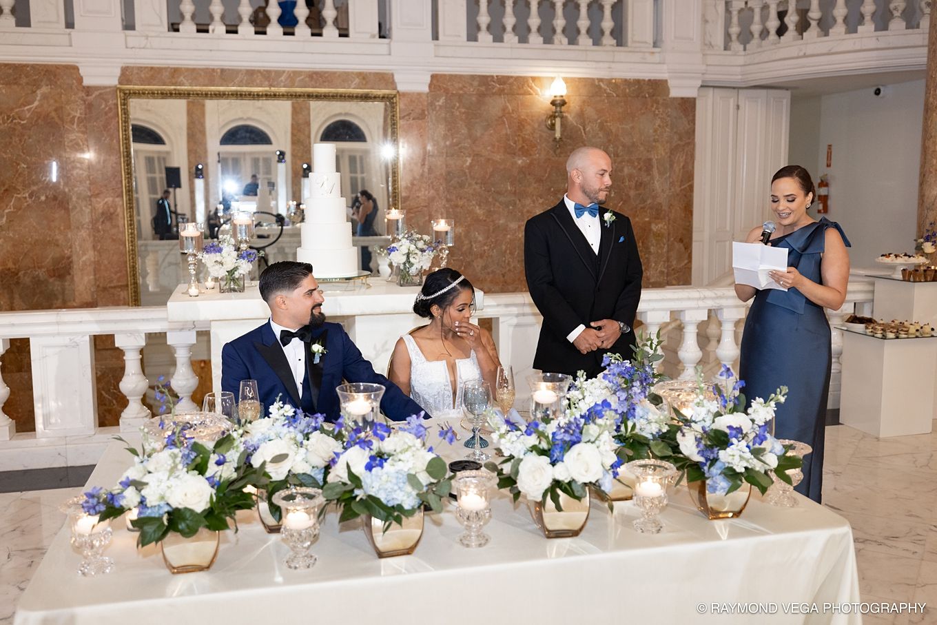 Wedding reception toast with bride and groom seated at floral-decorated head table while guests deliver speeches in elegant venue.