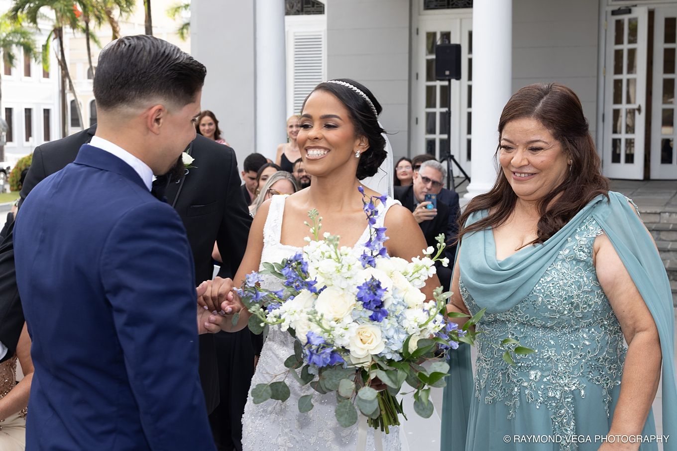 Bride in white gown holds blue and white bouquet, smiling with groom and guest at outdoor wedding ceremony.