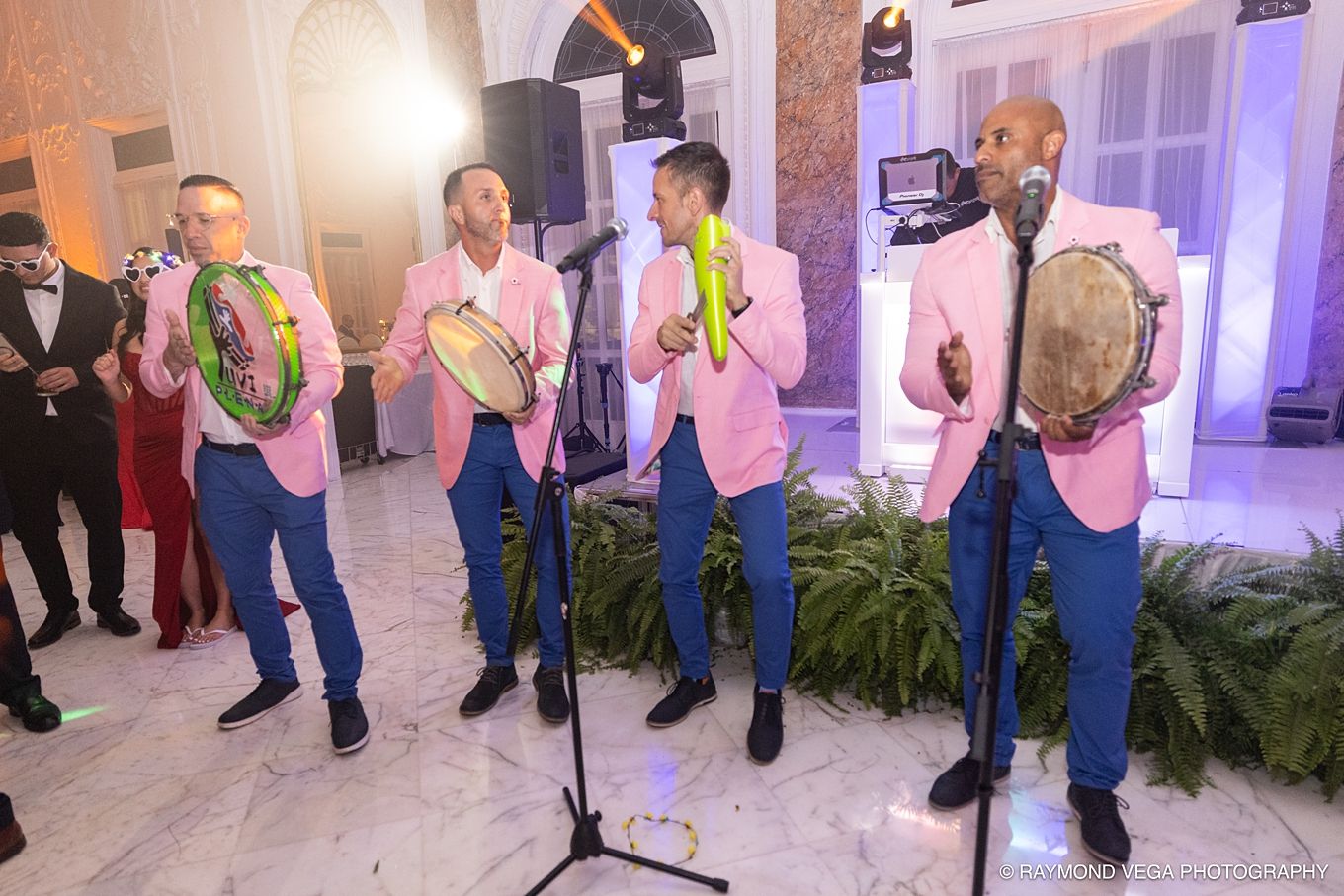 Four men in pink blazers and blue pants play tambourines at an elegant indoor event. PLENA EN PUERTO RICO