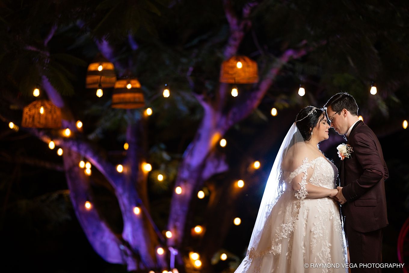 Romantic wedding couple sharing a kiss under glowing string lights and purple-lit trees at an enchanting outdoor night ceremony.
