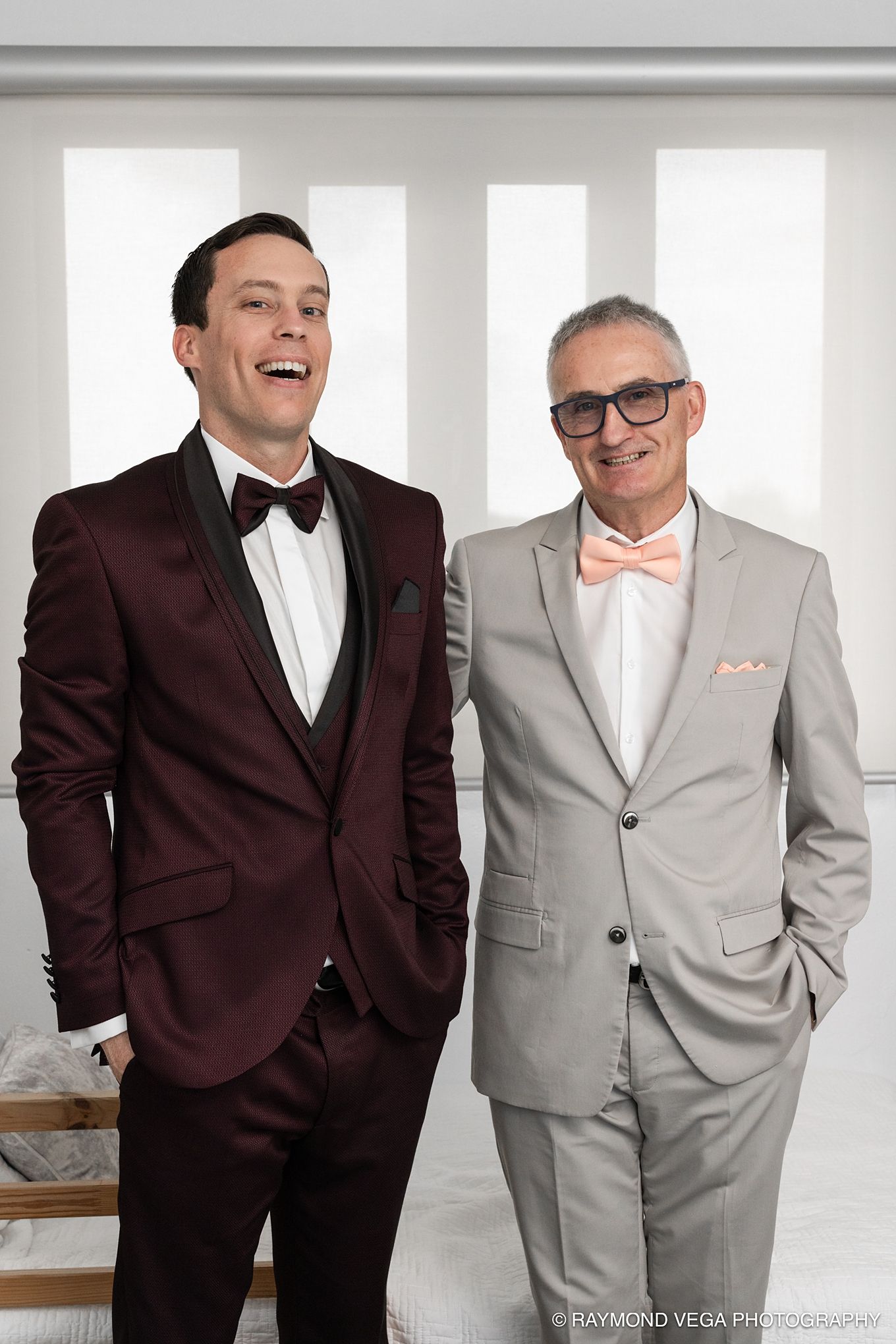 Two men in formal suits with bow ties smiling together at a wedding, one in burgundy tuxedo, one in gray suit.