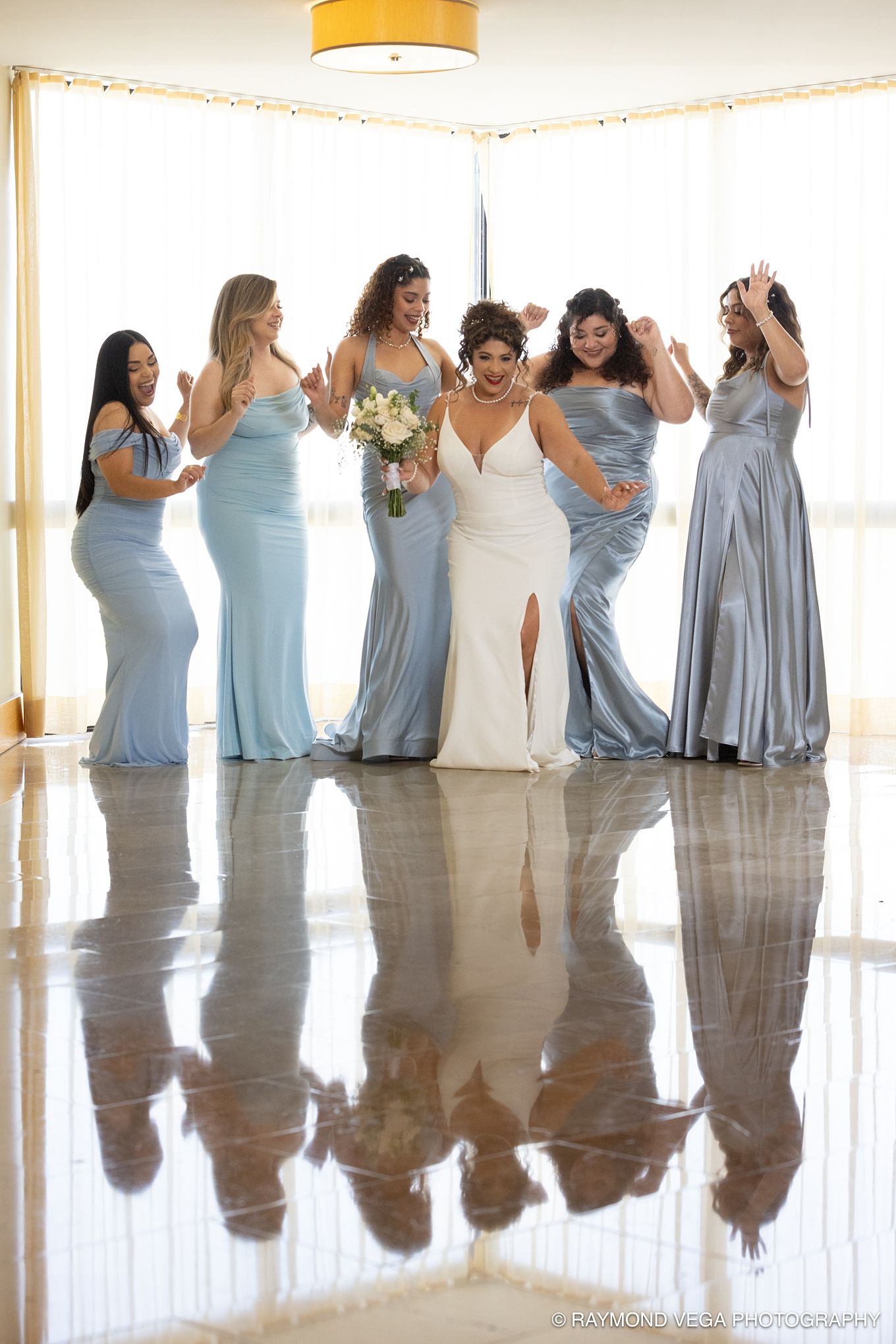 Bride in white gown poses with six bridesmaids in blue satin dresses, all celebrating on a reflective floor.