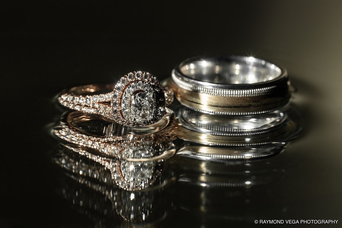 Elegant diamond engagement ring and wedding bands reflected on a dark surface, photographed by Raymond Vega Photography.