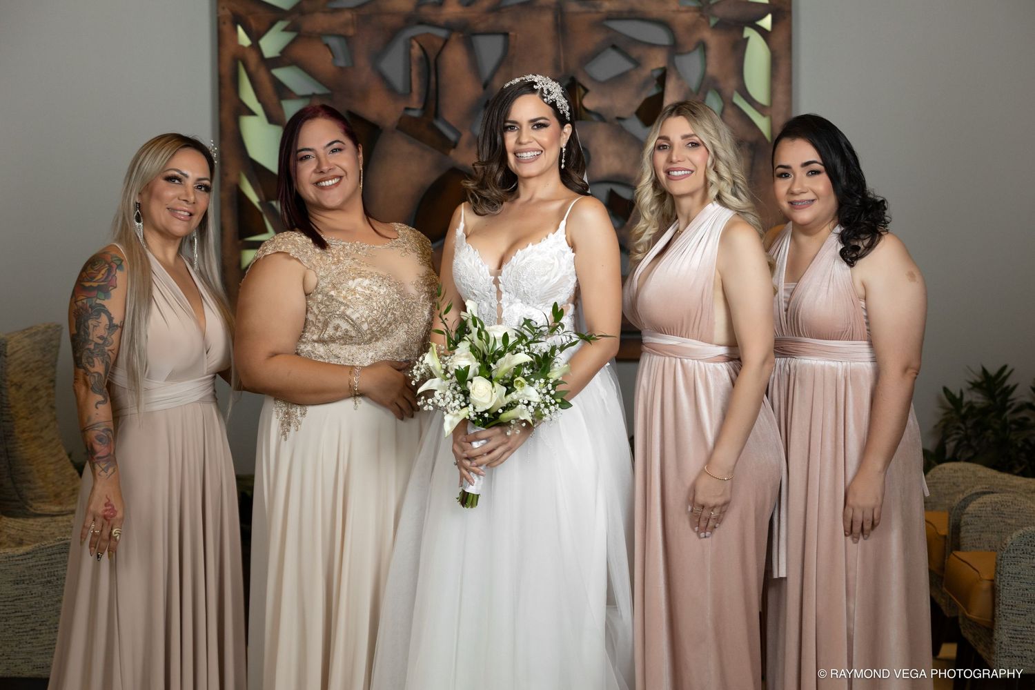 Bride in white gown holding bouquet poses with four bridesmaids in blush pink dresses at a wedding.