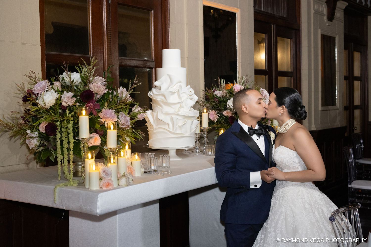 Newlyweds share a kiss beside a white wedding cake adorned with florals and candles at an elegant reception venue.