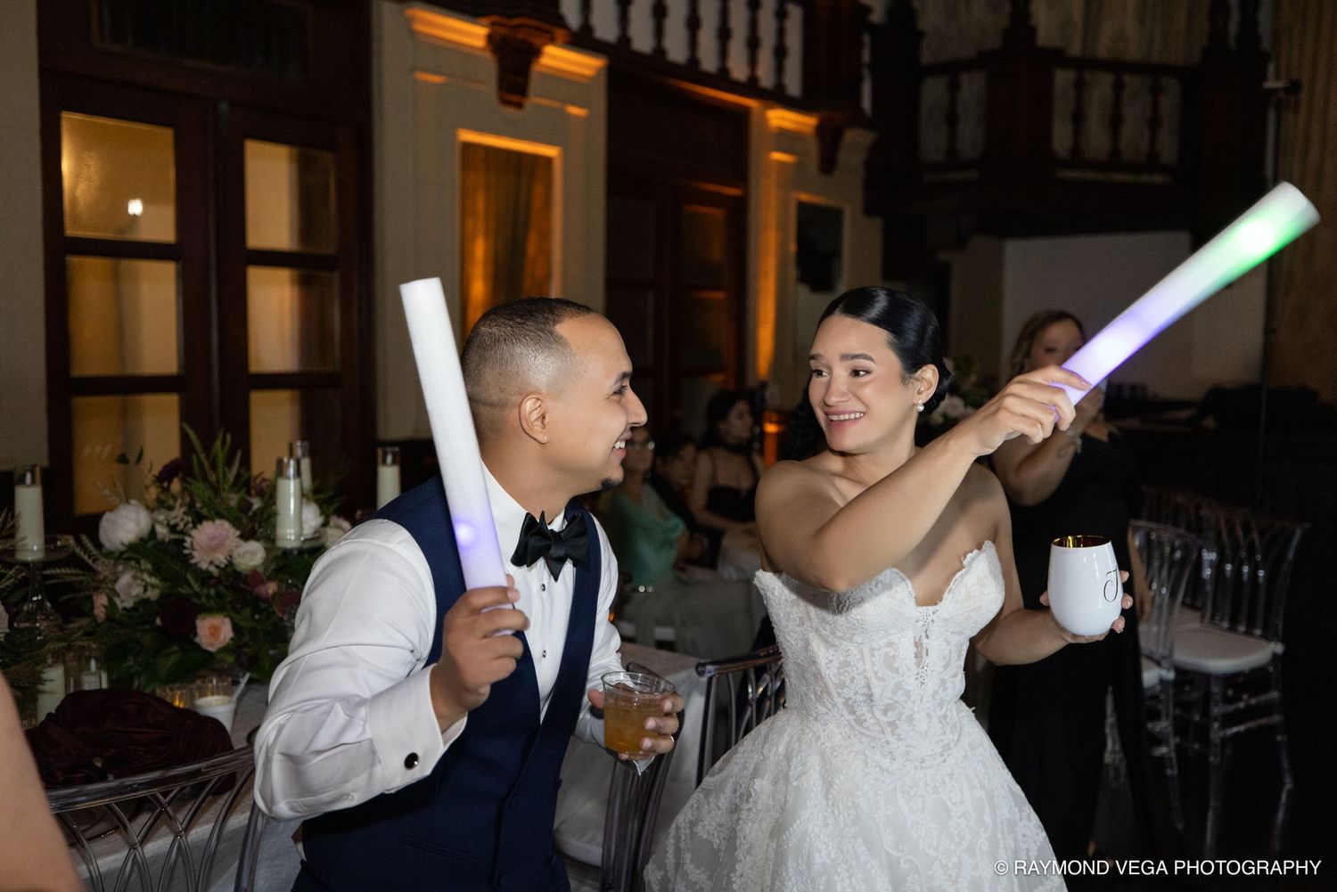 Bride and groom joyfully holding glowing light sticks at their wedding reception in an elegant venue.