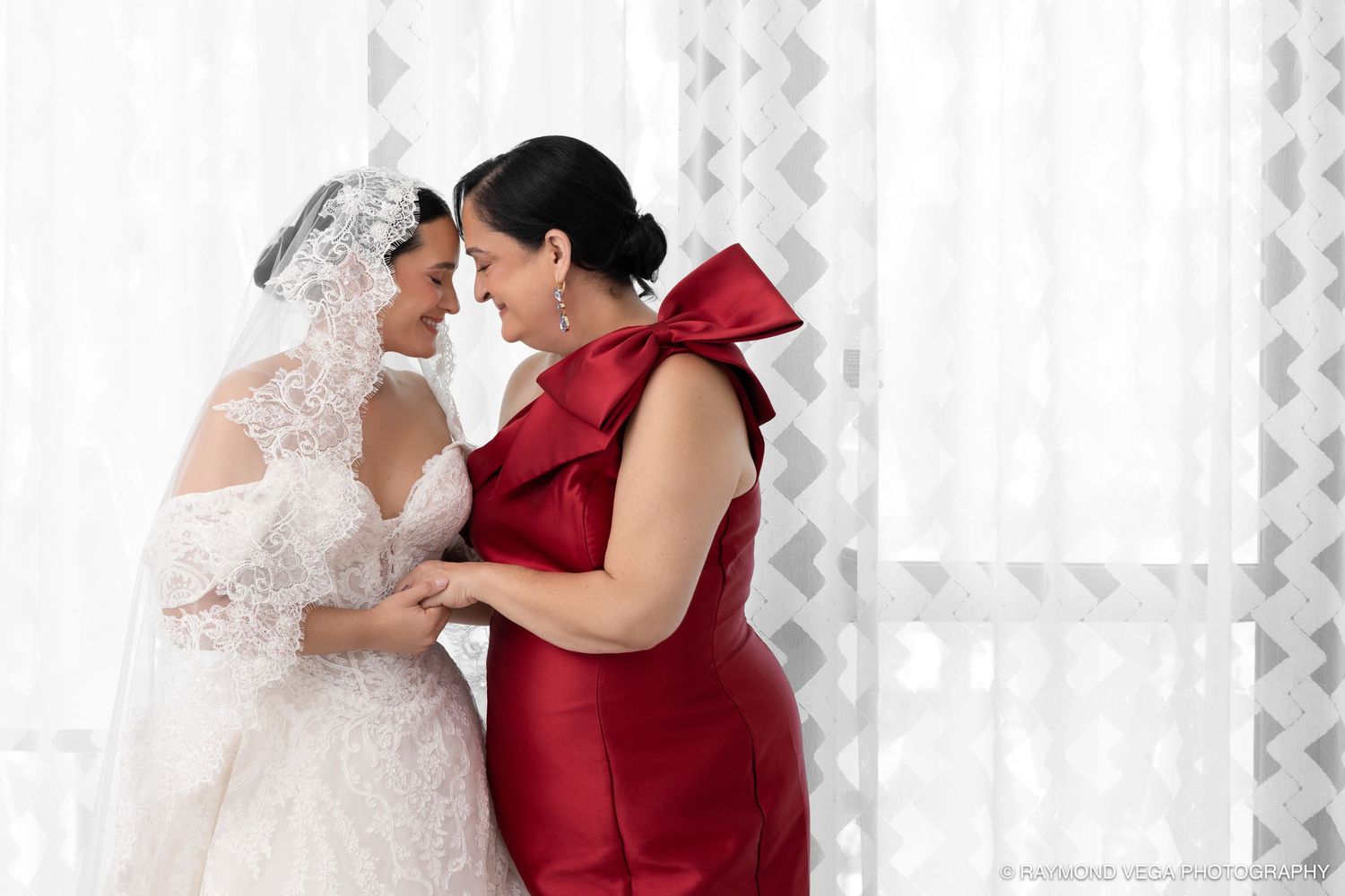 Bride in white lace gown and veil shares tender moment with woman in red bow dress near sheer curtains.