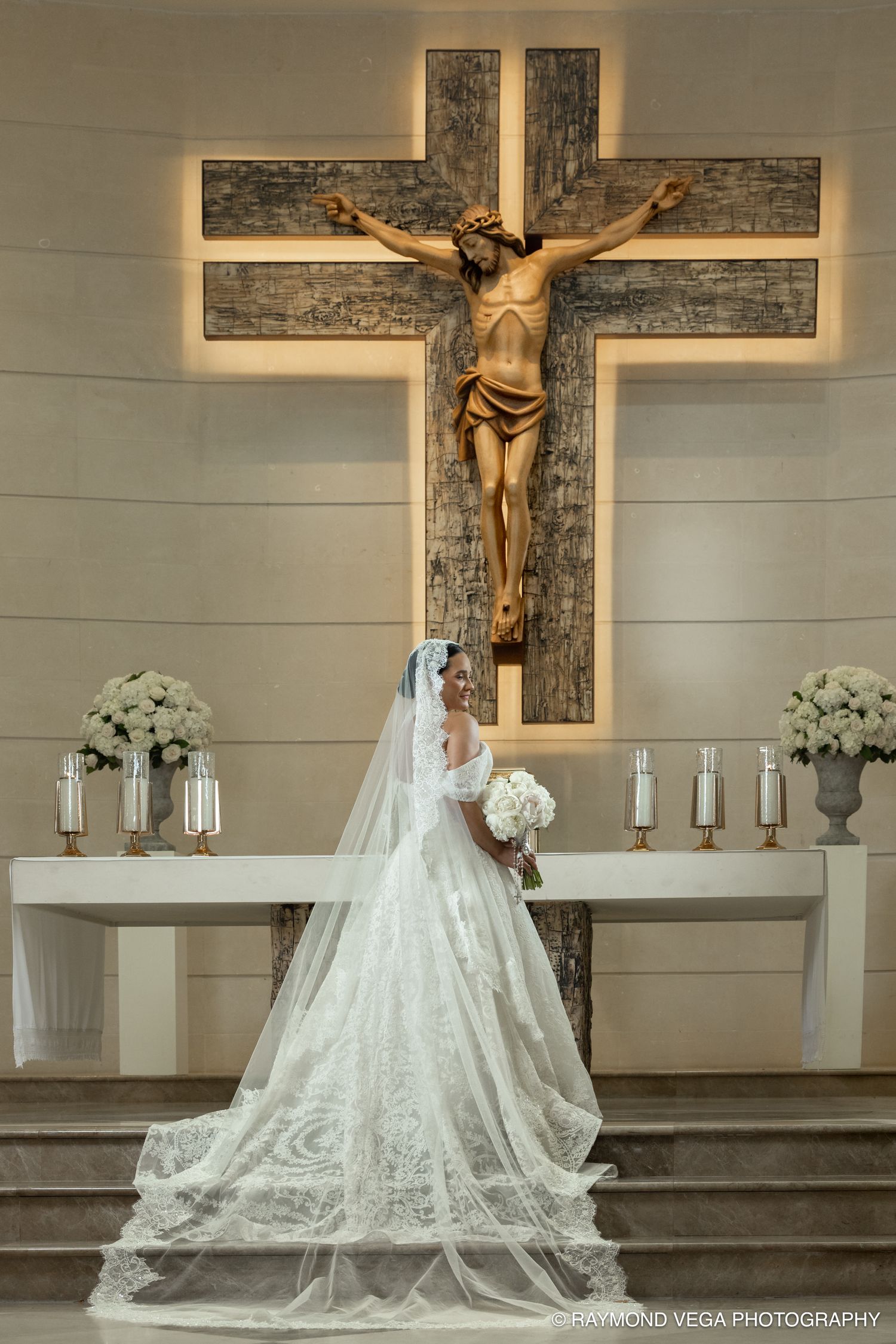 Bride in white lace gown and veil stands before crucifix altar holding bouquet in elegant church wedding ceremony.