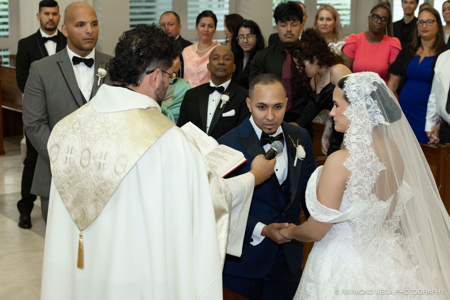 A couple exchanges wedding vows at a church altar with a priest officiating, surrounded by guests in formal attire.
