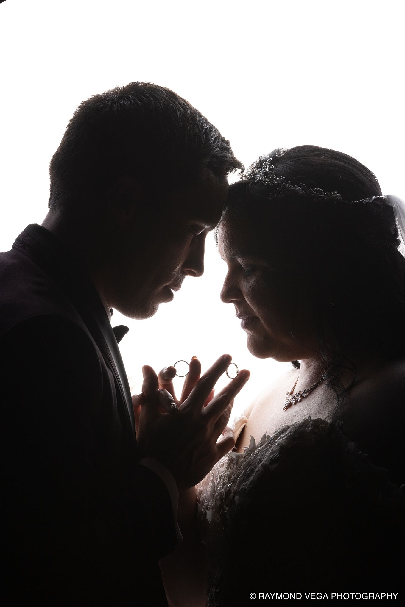 Silhouette of a bride and groom holding wedding rings together, captured by Raymond Vega Photography.