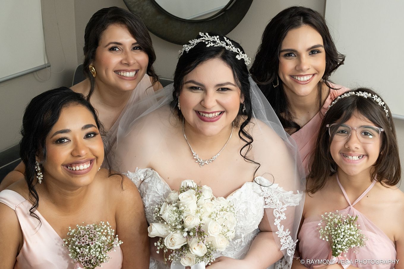 Bride in white gown and veil poses with four bridesmaids in pink dresses, all smiling and holding white floral bouquets.
