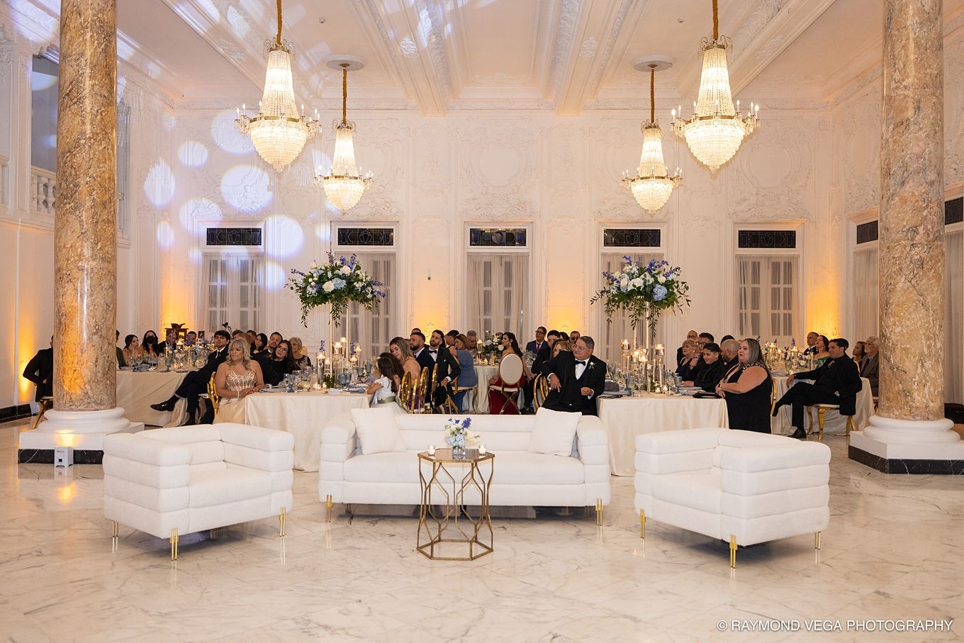 Elegant wedding reception in a grand marble ballroom with white lounge furniture, crystal chandeliers, and floral centerpieces.