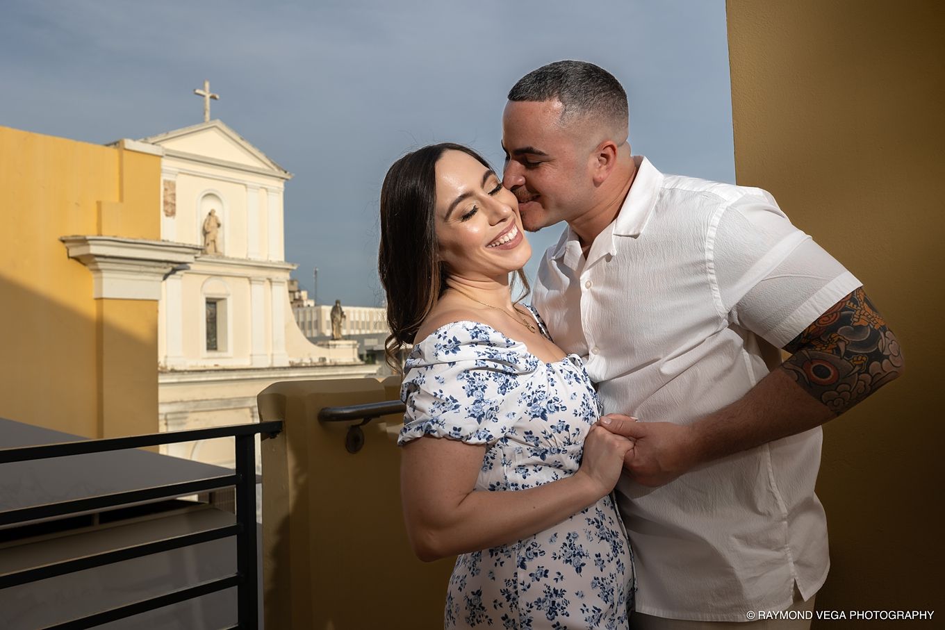 Couple sharing a romantic embrace on a balcony with a historic church and blue sky in the background. - Love Story
