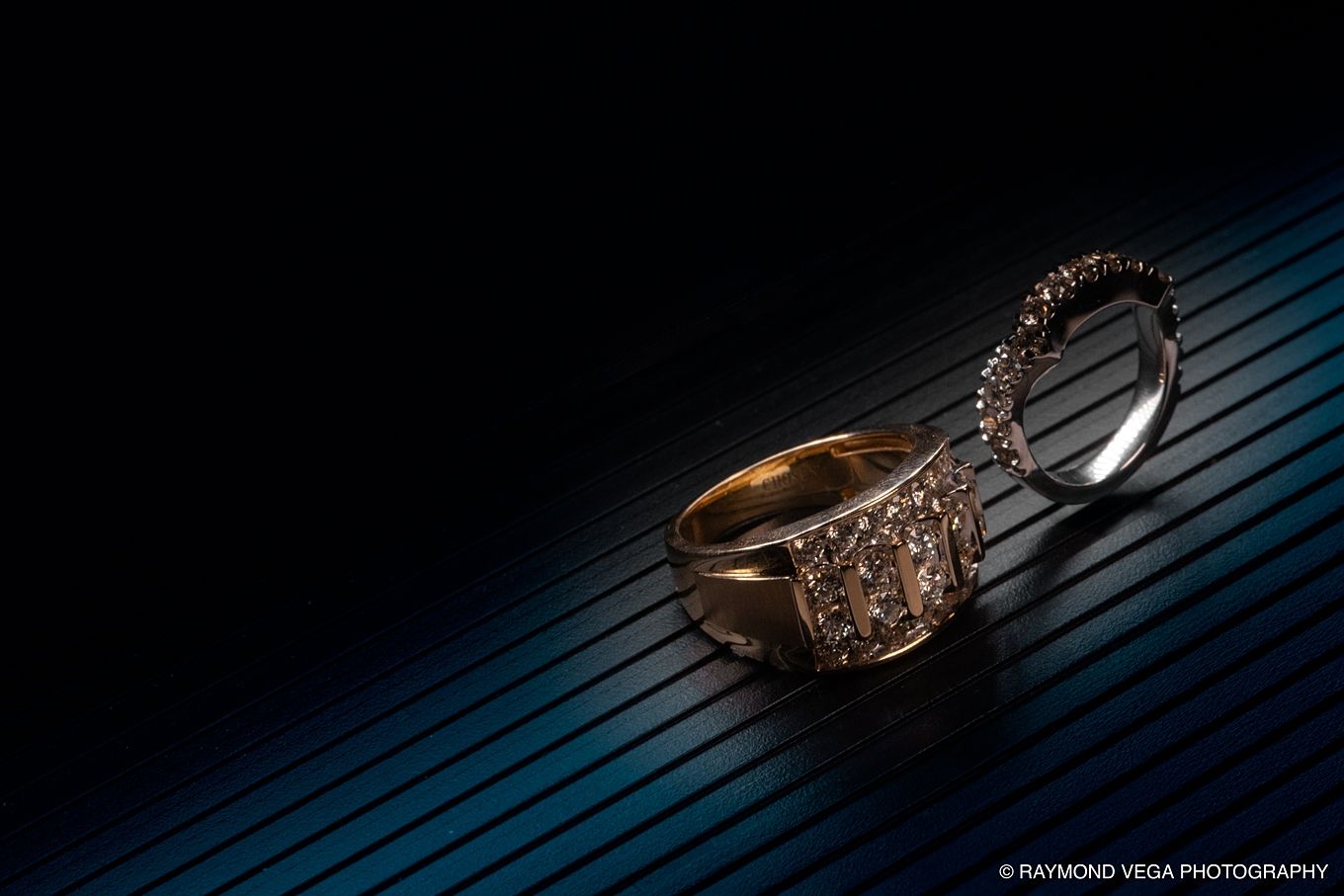 Gold diamond engagement ring and wedding band displayed on dark blue striped surface by Raymond Vega Photography.