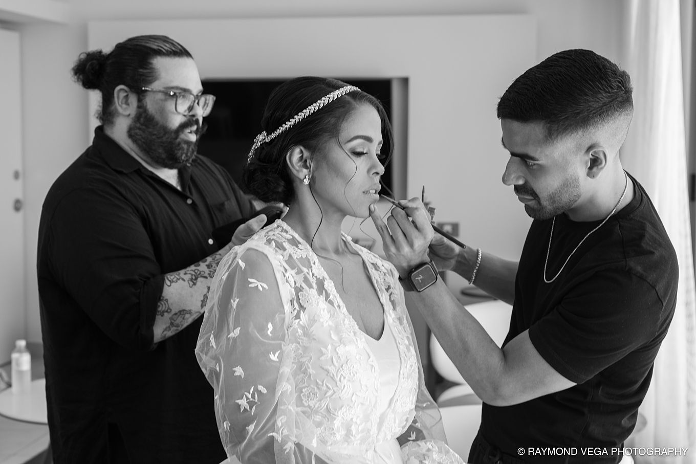Bride getting makeup applied by two stylists in a lace wedding gown, black and white wedding preparation photo.