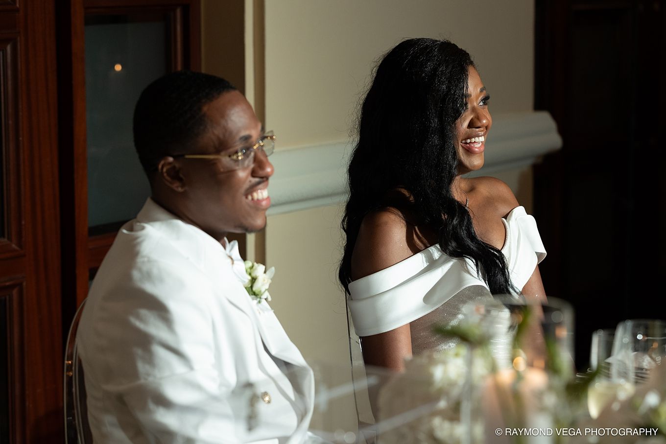 A joyful couple in white wedding attire laughing together at their reception dinner table.