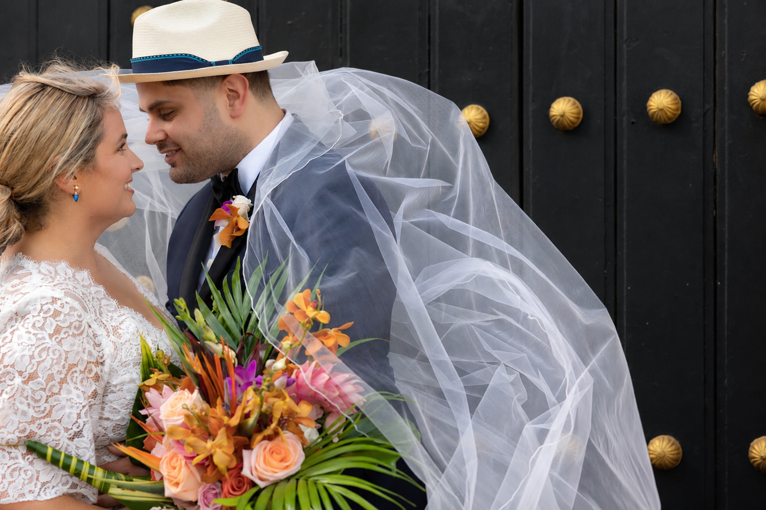 Bride and groom share romantic moment under veil, holding tropical bouquet of orange and pink flowers by ornate black door.