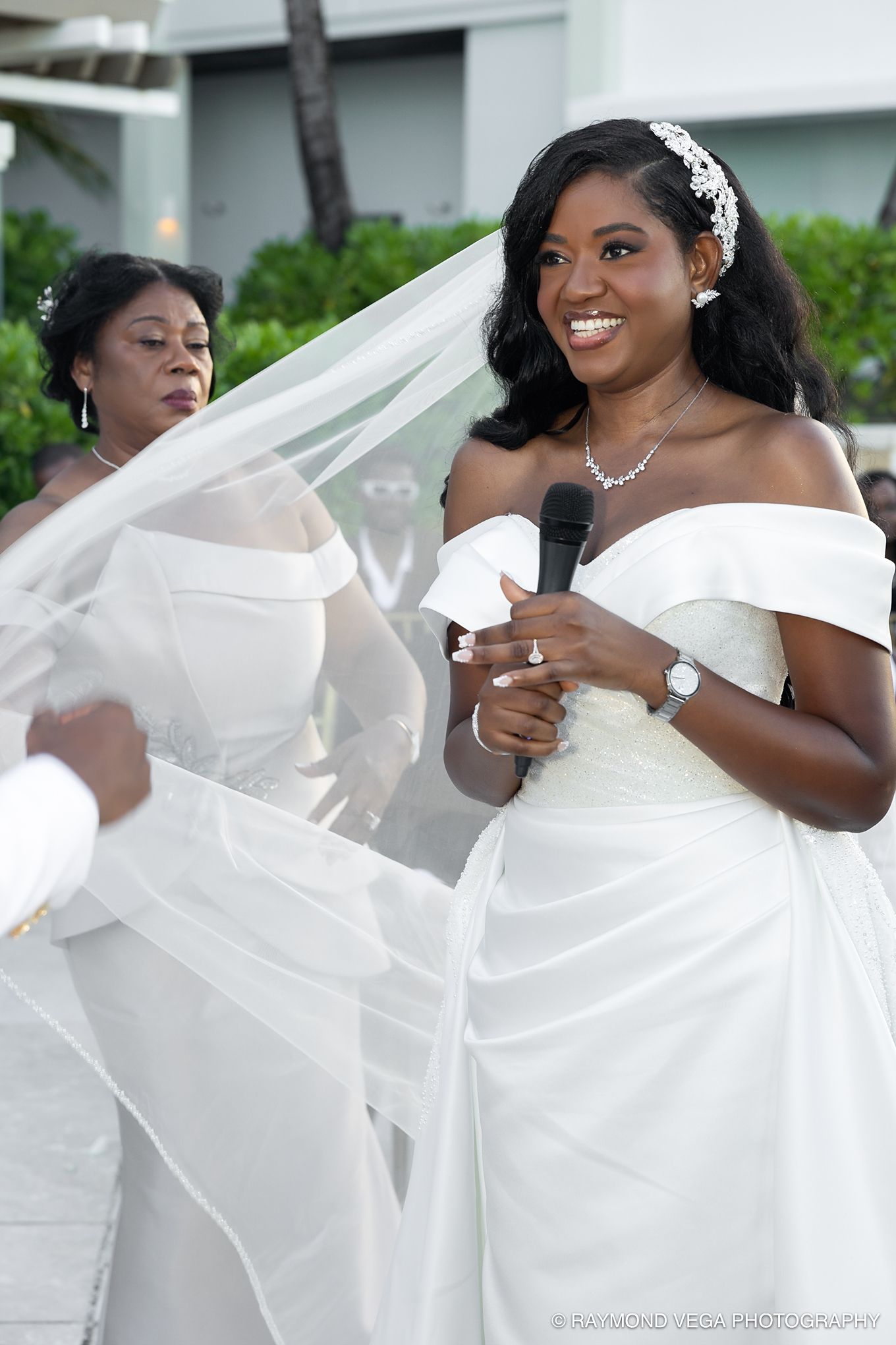 Smiling bride in off-shoulder white gown holds microphone at outdoor wedding ceremony with veil flowing.