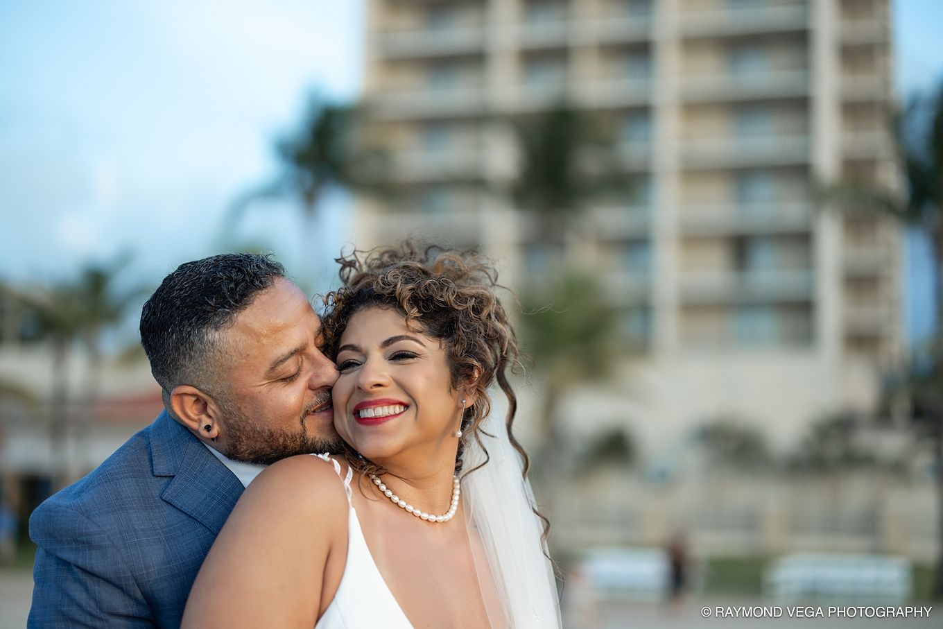 Bride and groom share a romantic moment outdoors, with the groom kissing her cheek as she smiles in her wedding veil.