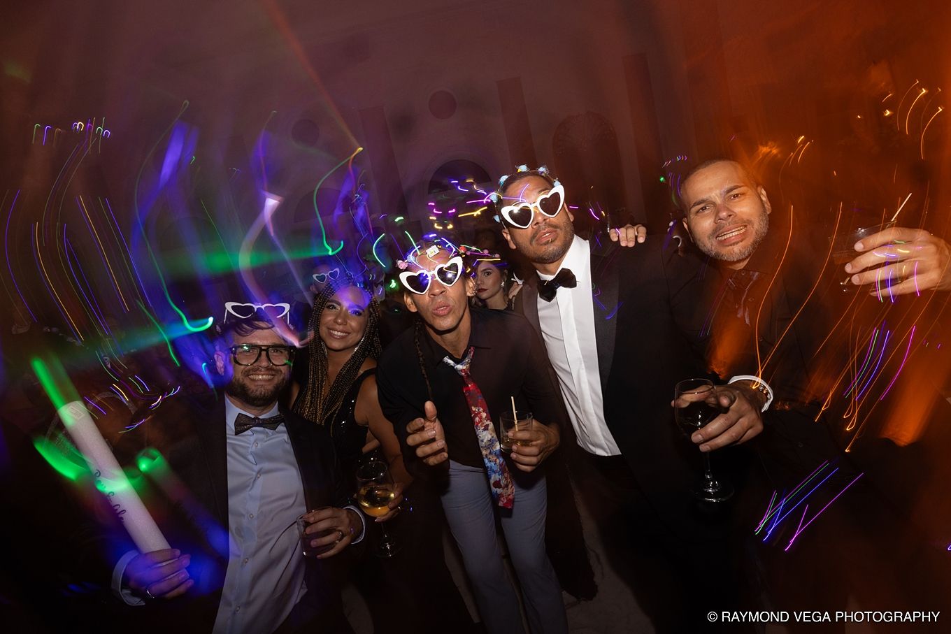 Group of friends posing at a party with glow sticks and fun glasses under colorful lights. Antiguo Casino Puerto Rico wedding