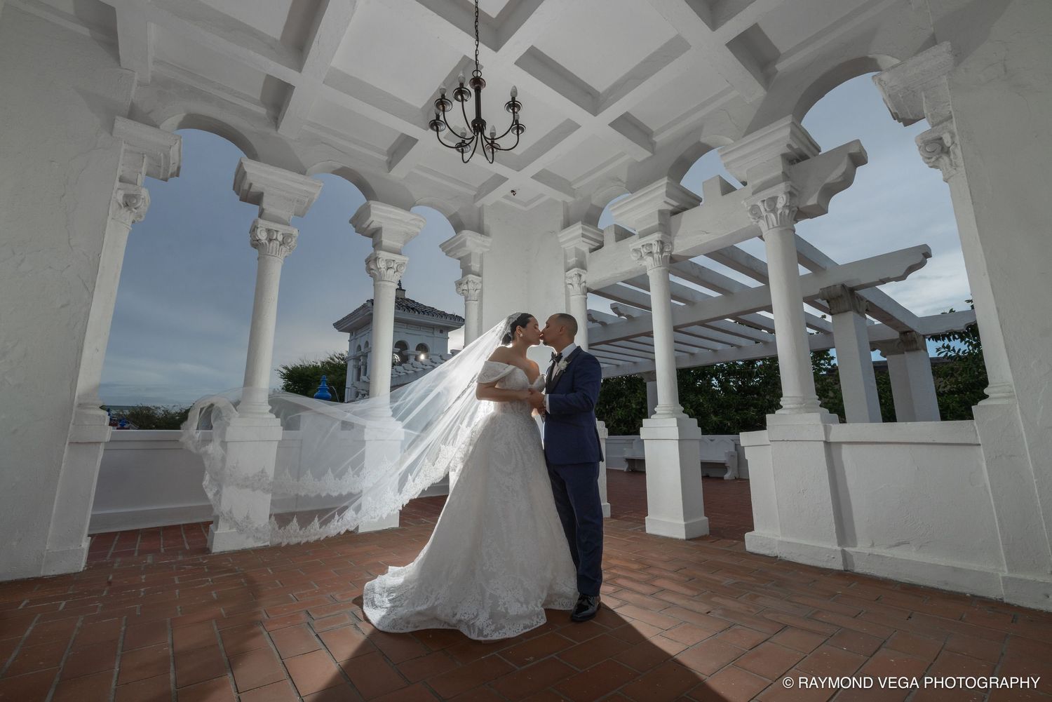 Bride and groom share a romantic kiss under white columns of an elegant wedding venue balcony.