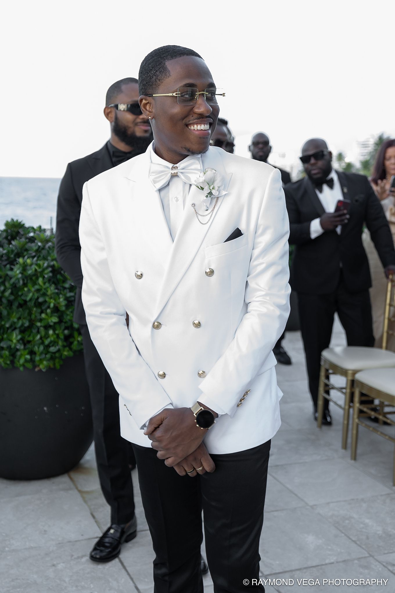 Smiling groom in white double-breasted tuxedo jacket with bow tie and boutonniere at outdoor wedding ceremony.