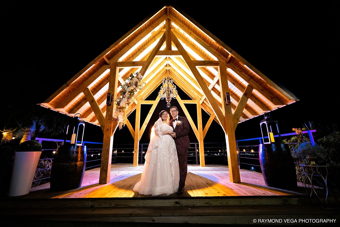 Bride and groom pose under illuminated wooden pavilion at night during outdoor wedding ceremony.