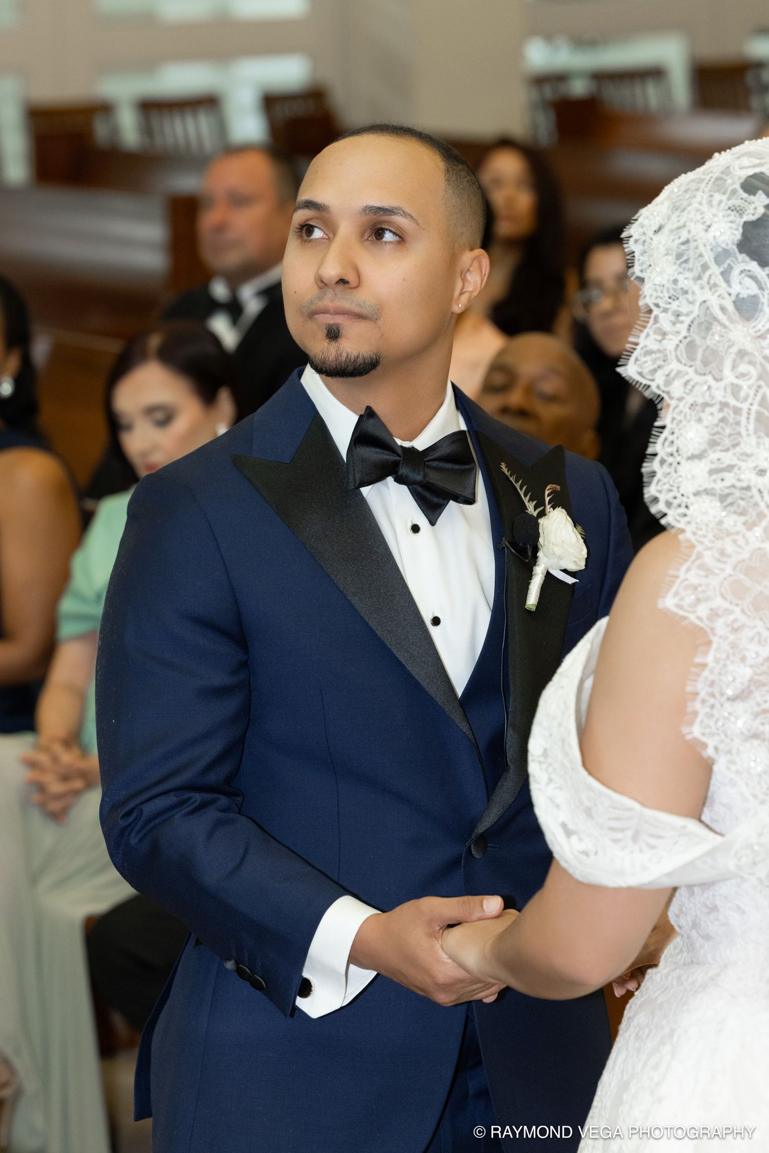 Groom in navy tuxedo holds bride's hands at wedding ceremony altar, looking up with emotional expression.