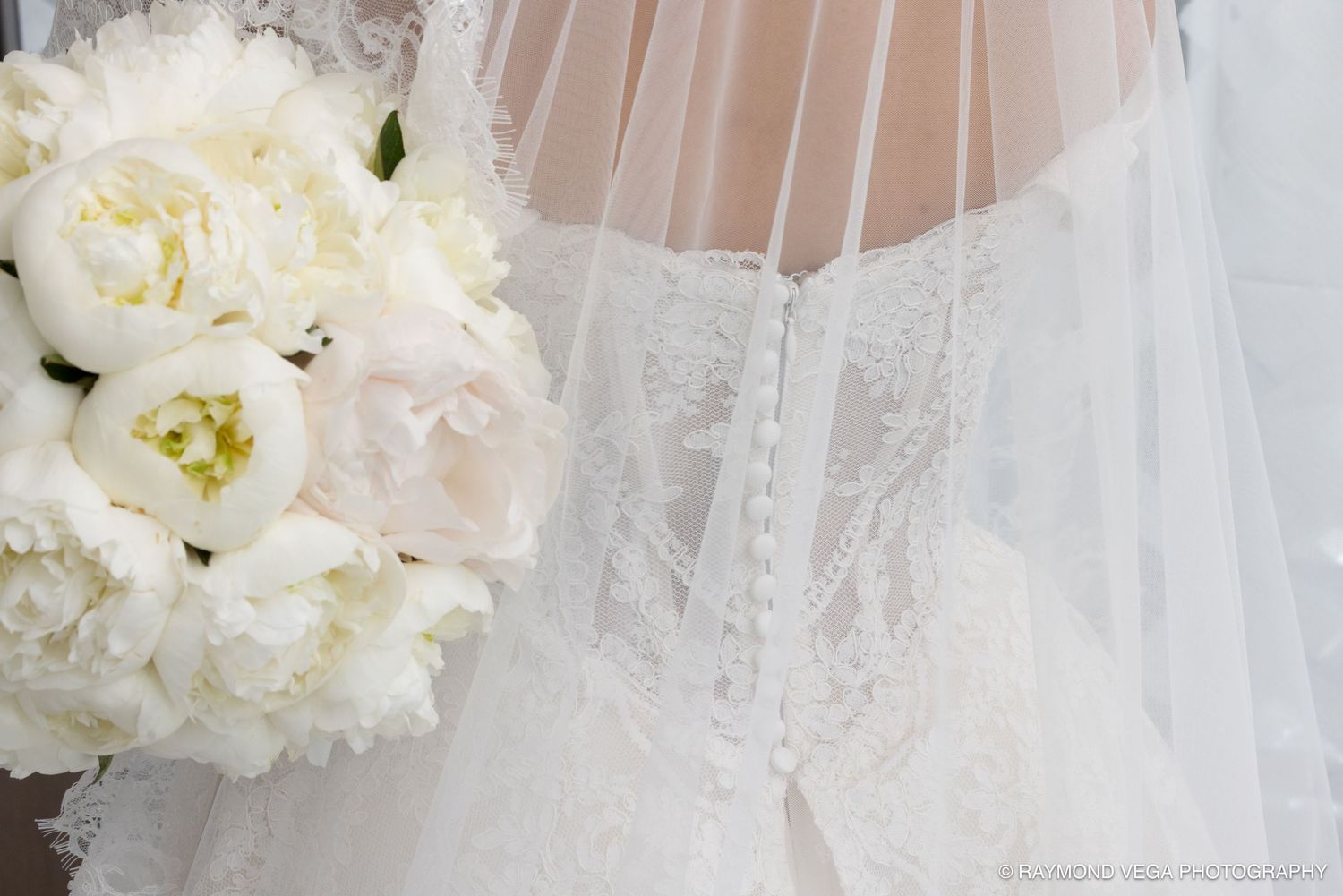 Close-up of a bride in a lace wedding dress holding a white peony bouquet, with veil draped over her back.