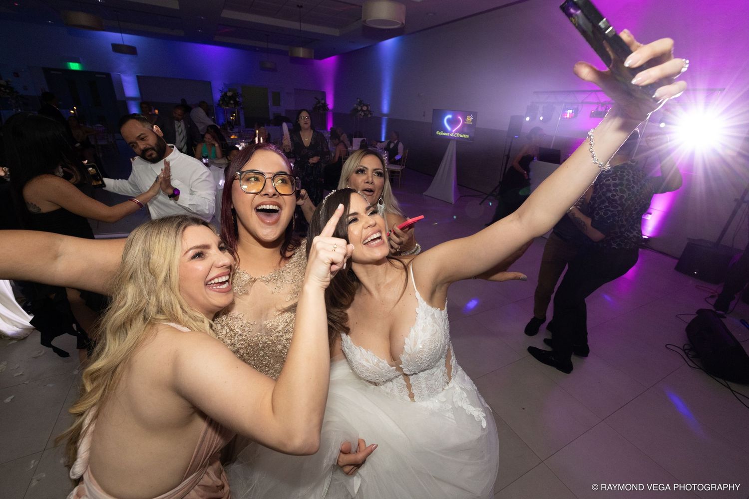 Bride and friends take a joyful selfie on the dance floor at a wedding reception with purple lighting.