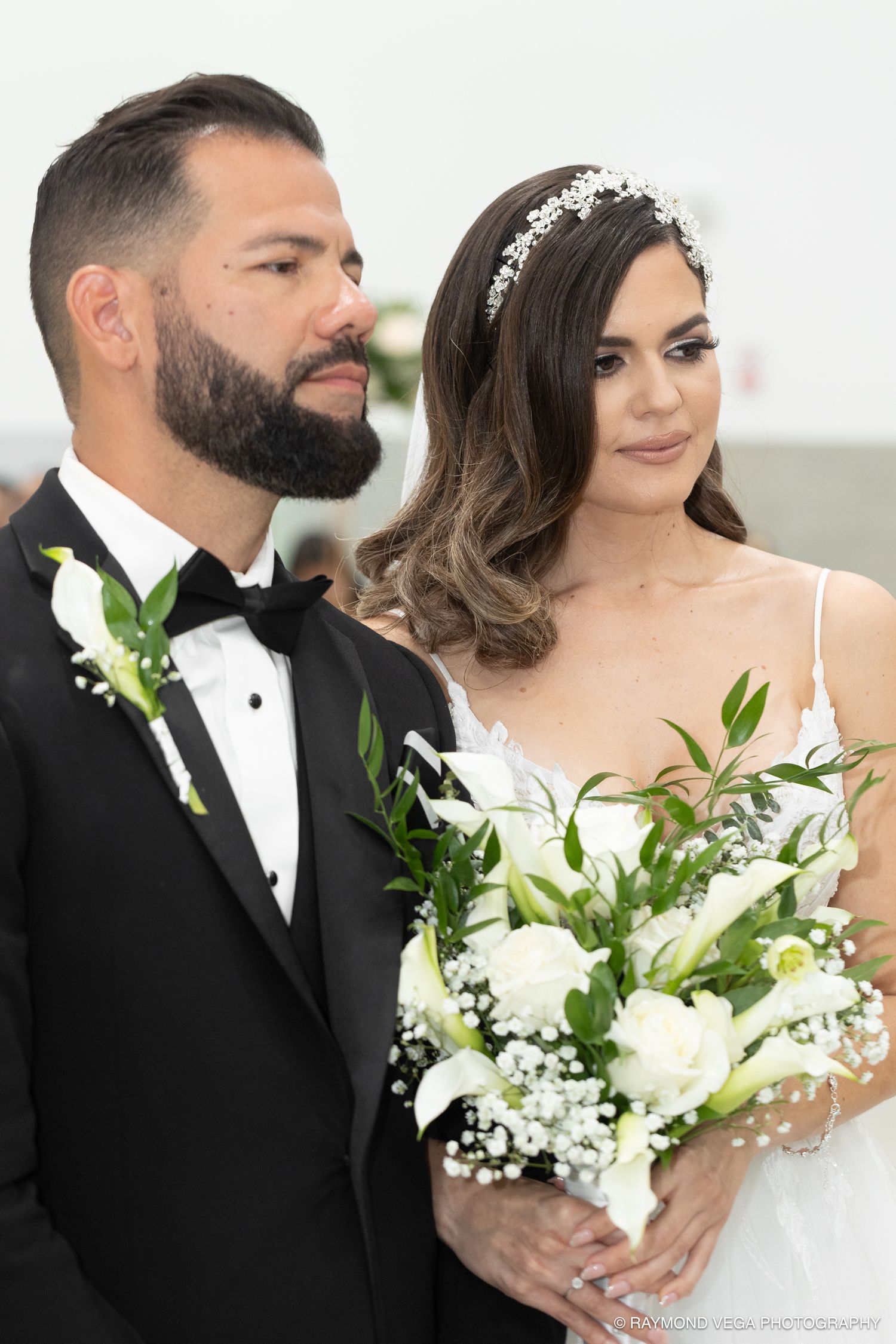 Bride holding white floral bouquet with groom in black tuxedo at elegant wedding ceremony.