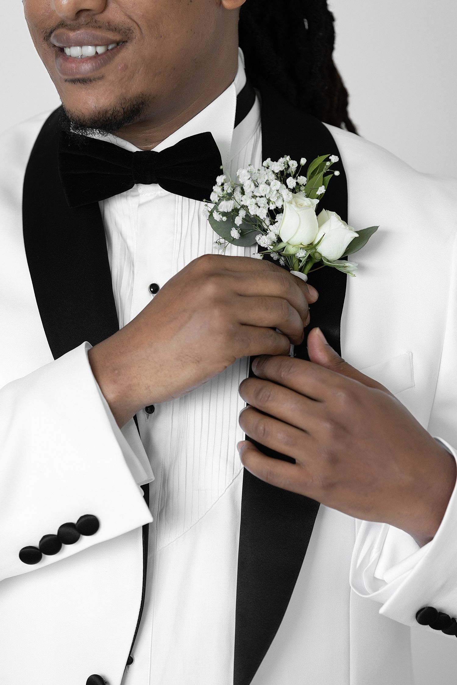 A smiling groom in a white tuxedo with black bow tie pins a white rose boutonniere to his lapel.