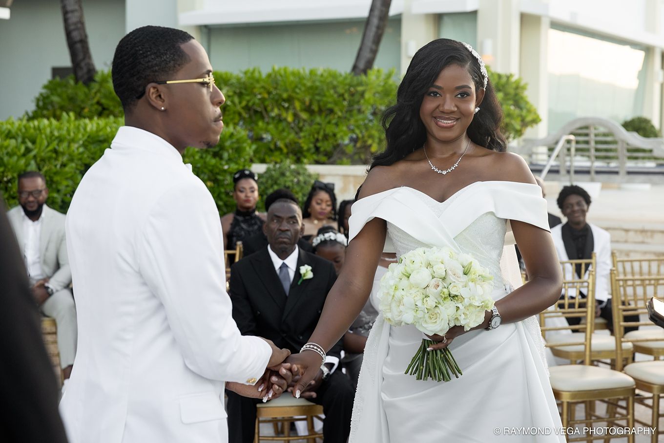 Bride and groom holding hands during outdoor wedding ceremony, bride wearing off-shoulder white gown holding white bouquet.