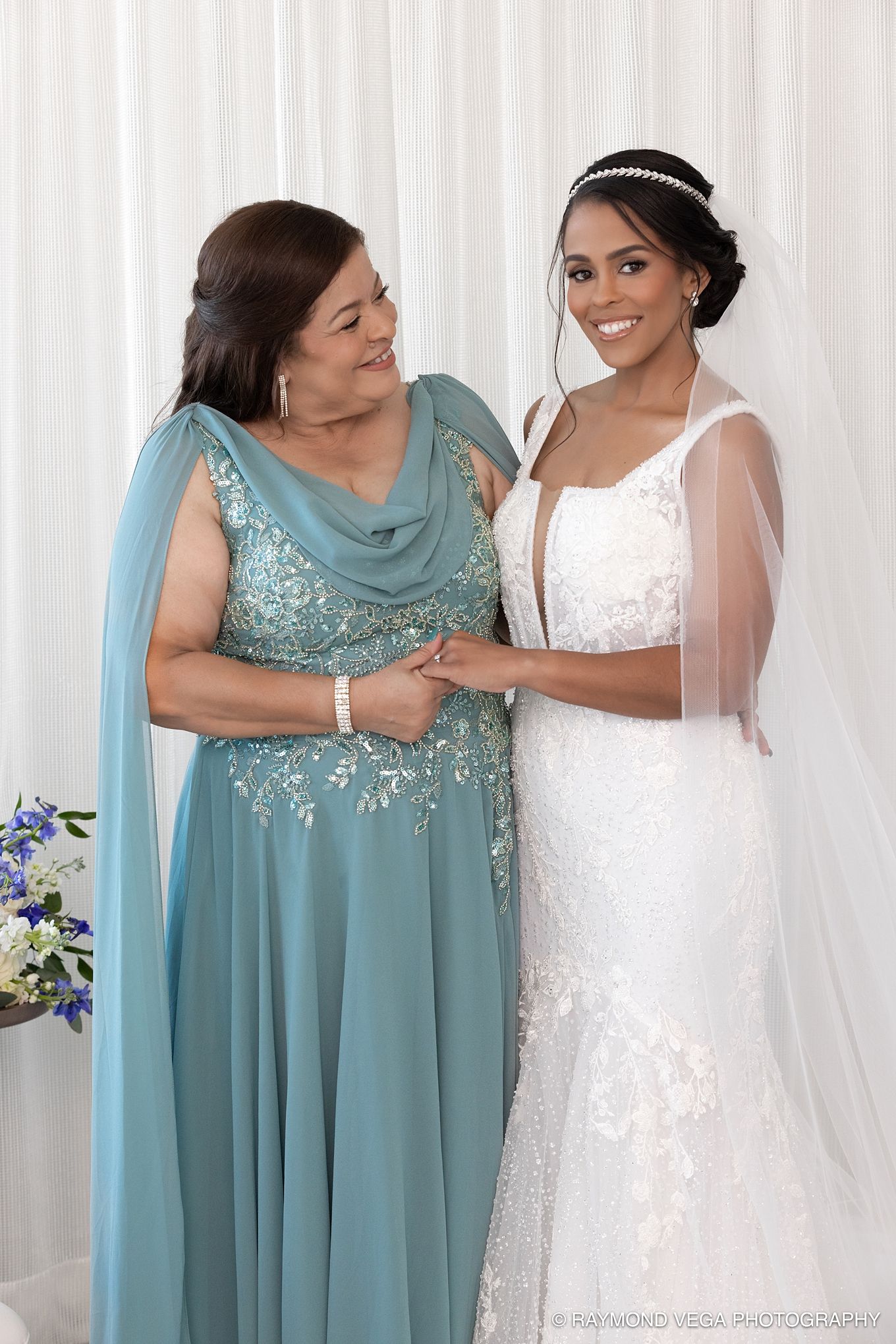 A bride in white lace gown and veil stands with an older woman in teal gown, holding hands and smiling warmly.
