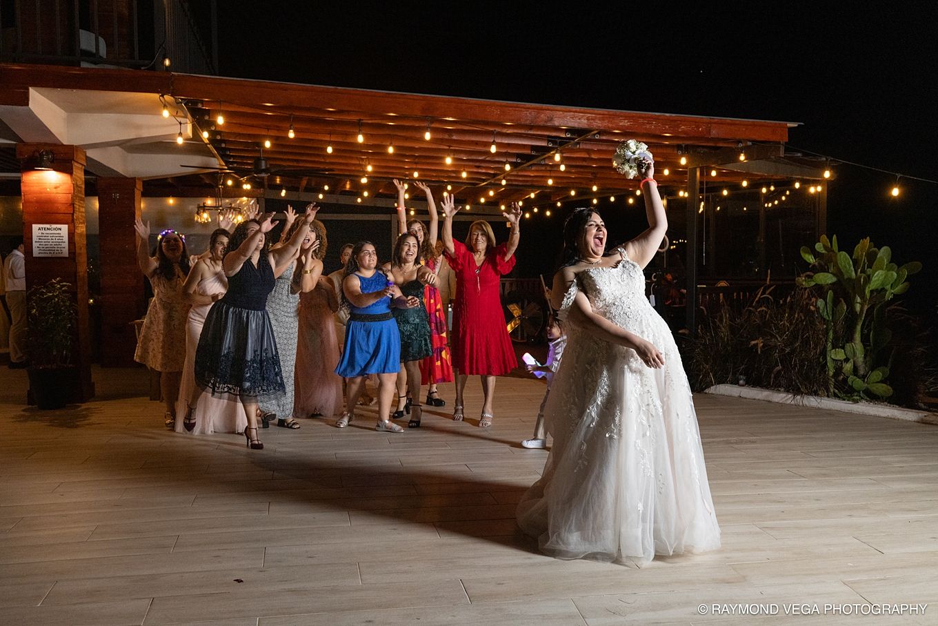Bride tossing bouquet to excited bridesmaids in colorful dresses at outdoor wedding reception at night.