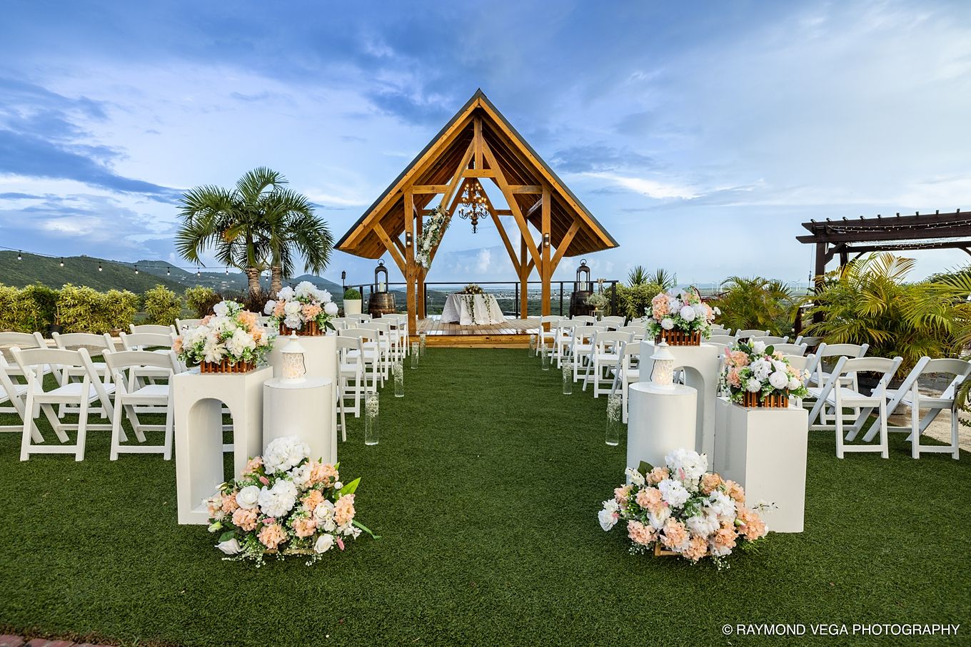 Elegant outdoor wedding ceremony setup with white chairs, floral arrangements, and a wooden gazebo overlooking tropical mountains.