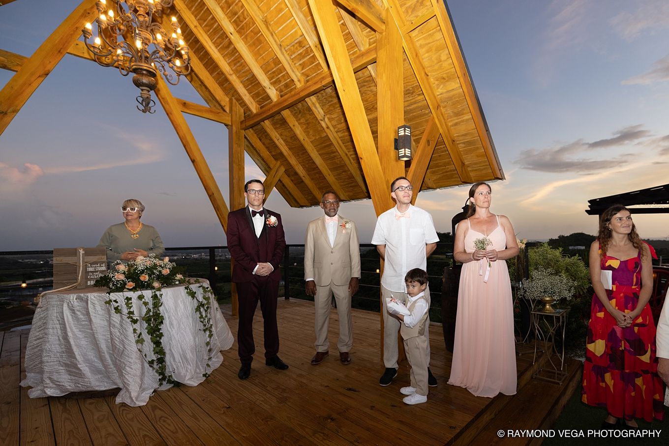 Outdoor wedding ceremony on a wooden deck at sunset with guests and wedding party gathered under a wooden pavilion.