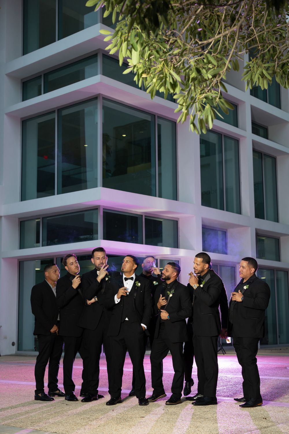 Groom and groomsmen in black tuxedos posing together at night outside a modern building with purple lighting.Raymond Vega Production