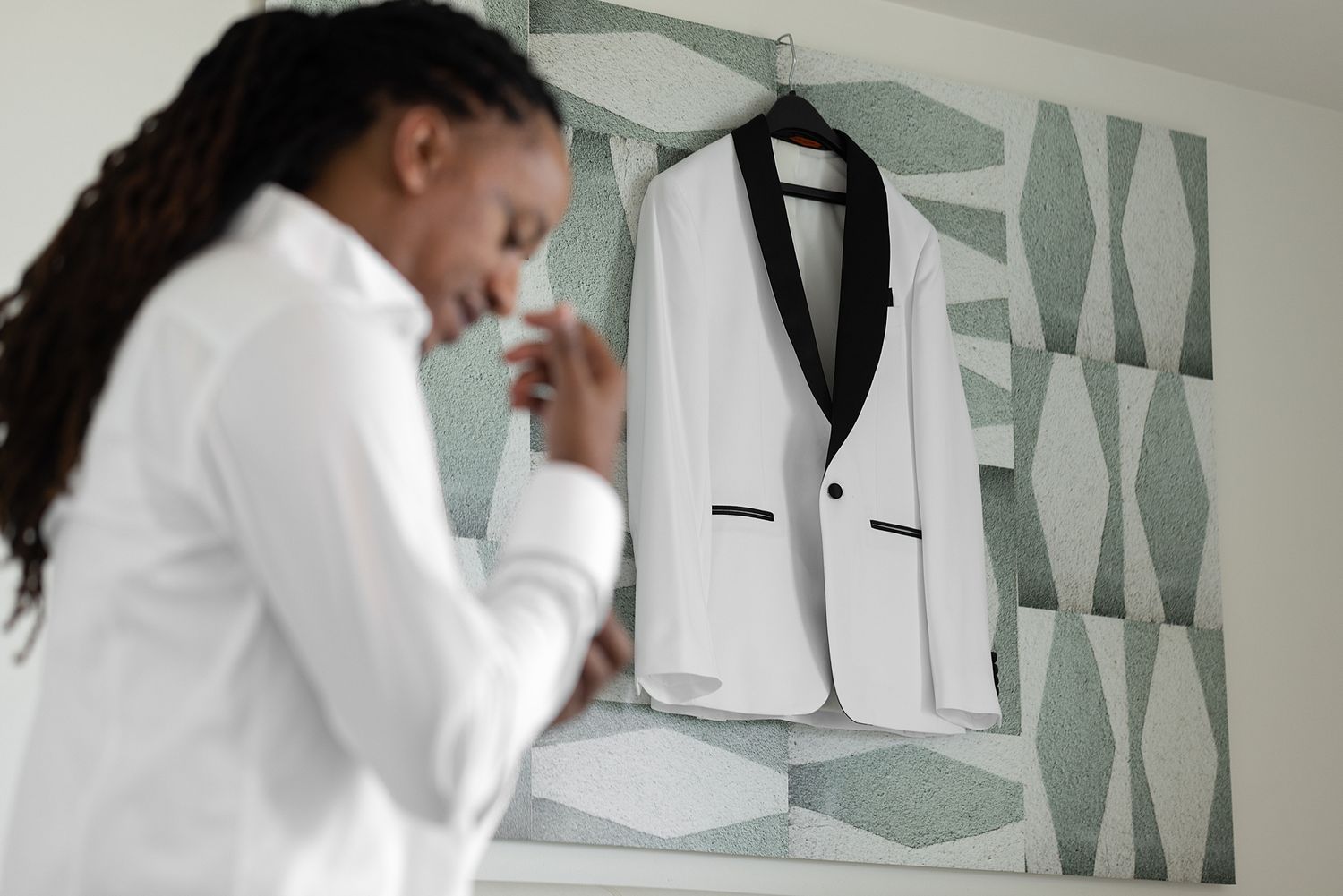 A man in a white shirt prepares beside a white tuxedo jacket with black lapels hanging on a patterned wall.