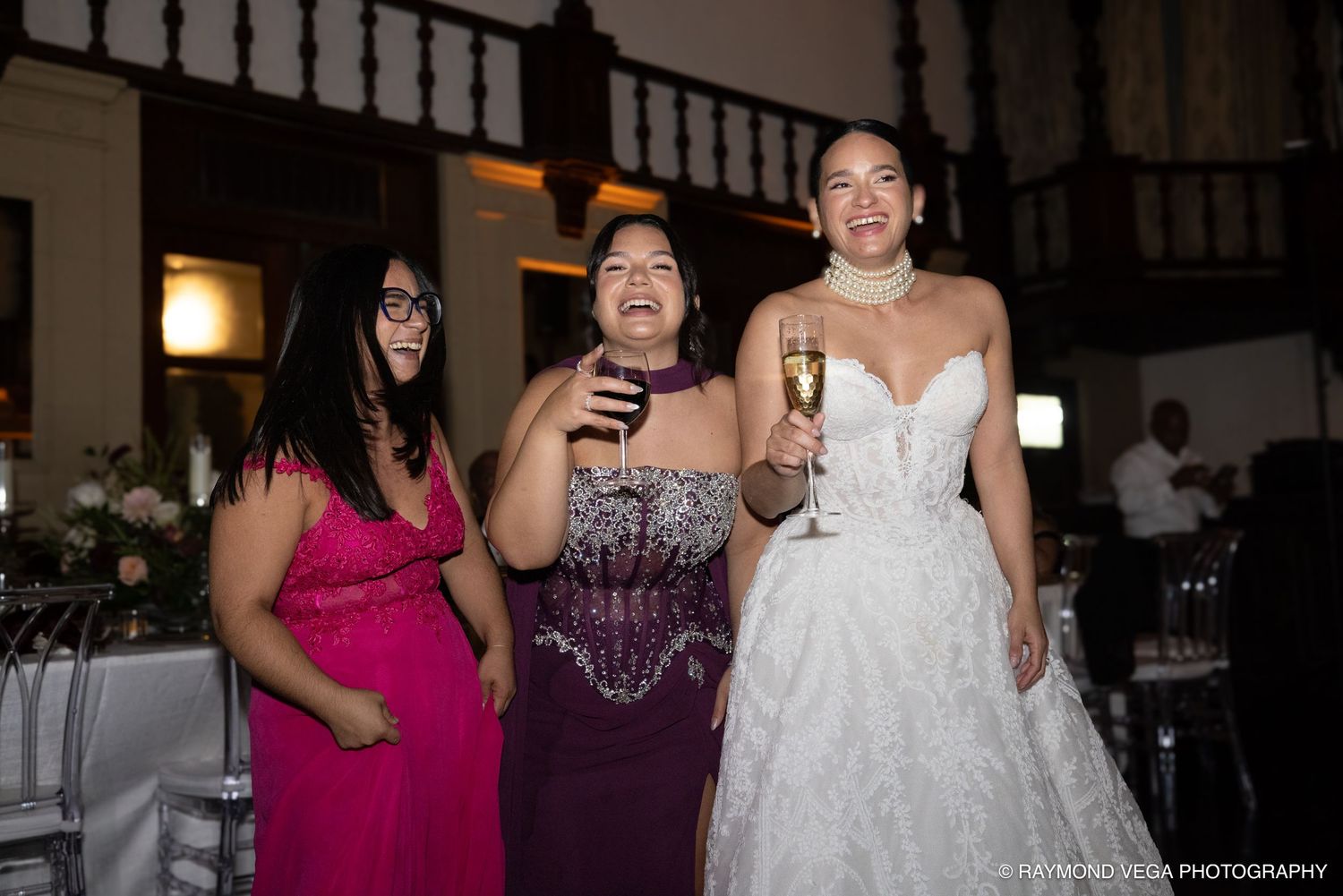 Bride in white gown laughing with two guests in pink and purple dresses at elegant wedding reception.