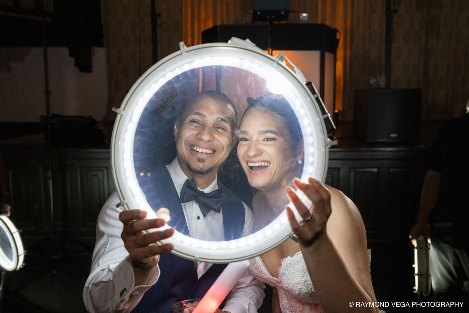 Bride and groom smiling through a lit circular drum frame at their wedding reception.