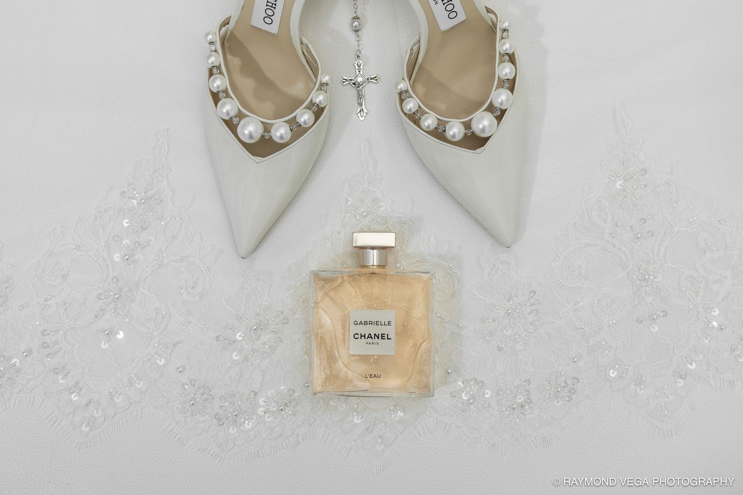 White pearl-embellished bridal heels beside a Chanel Gabrielle perfume bottle on a white lace background.
