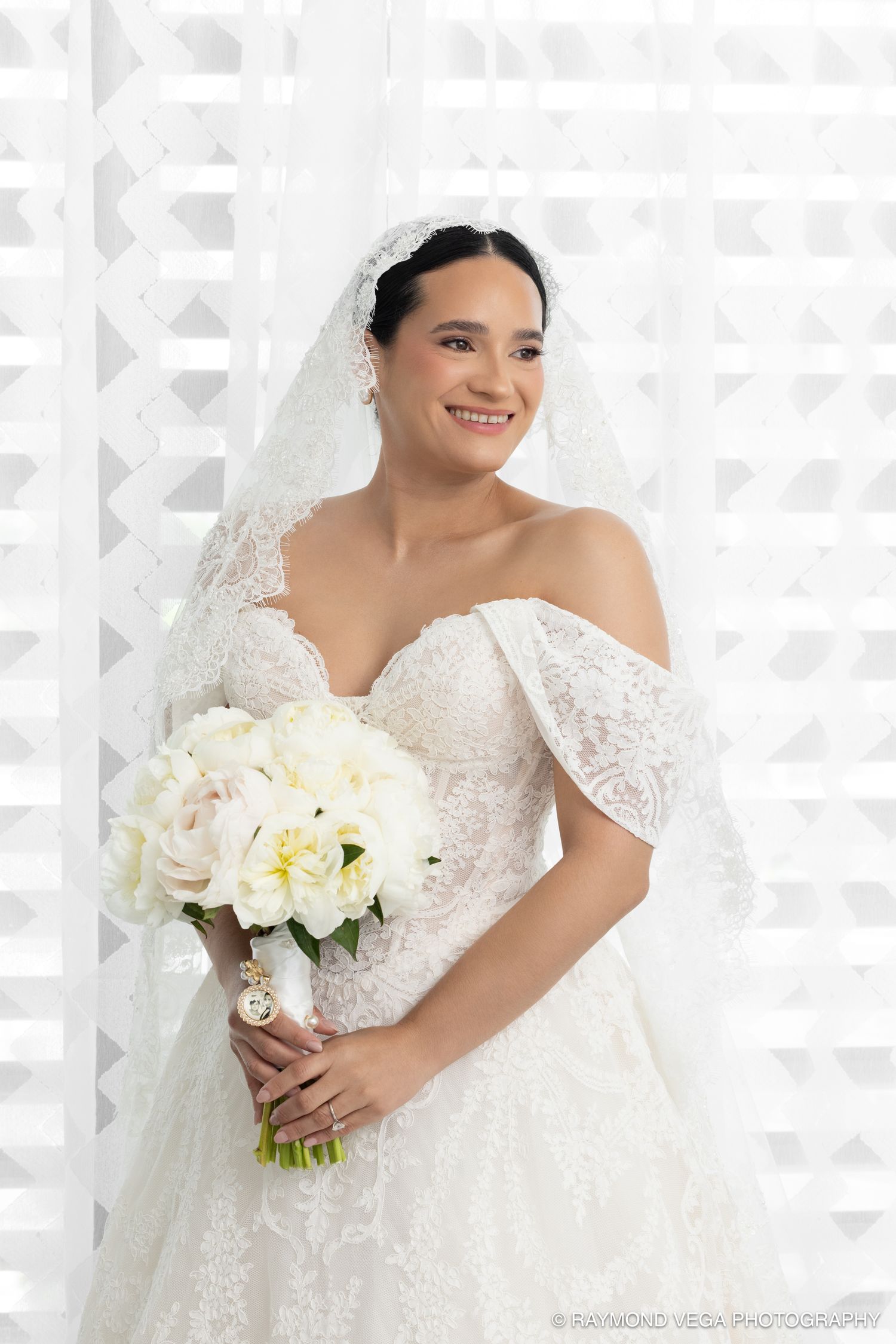 Smiling bride in off-shoulder lace wedding gown and veil holds white peony bouquet against geometric white backdrop.